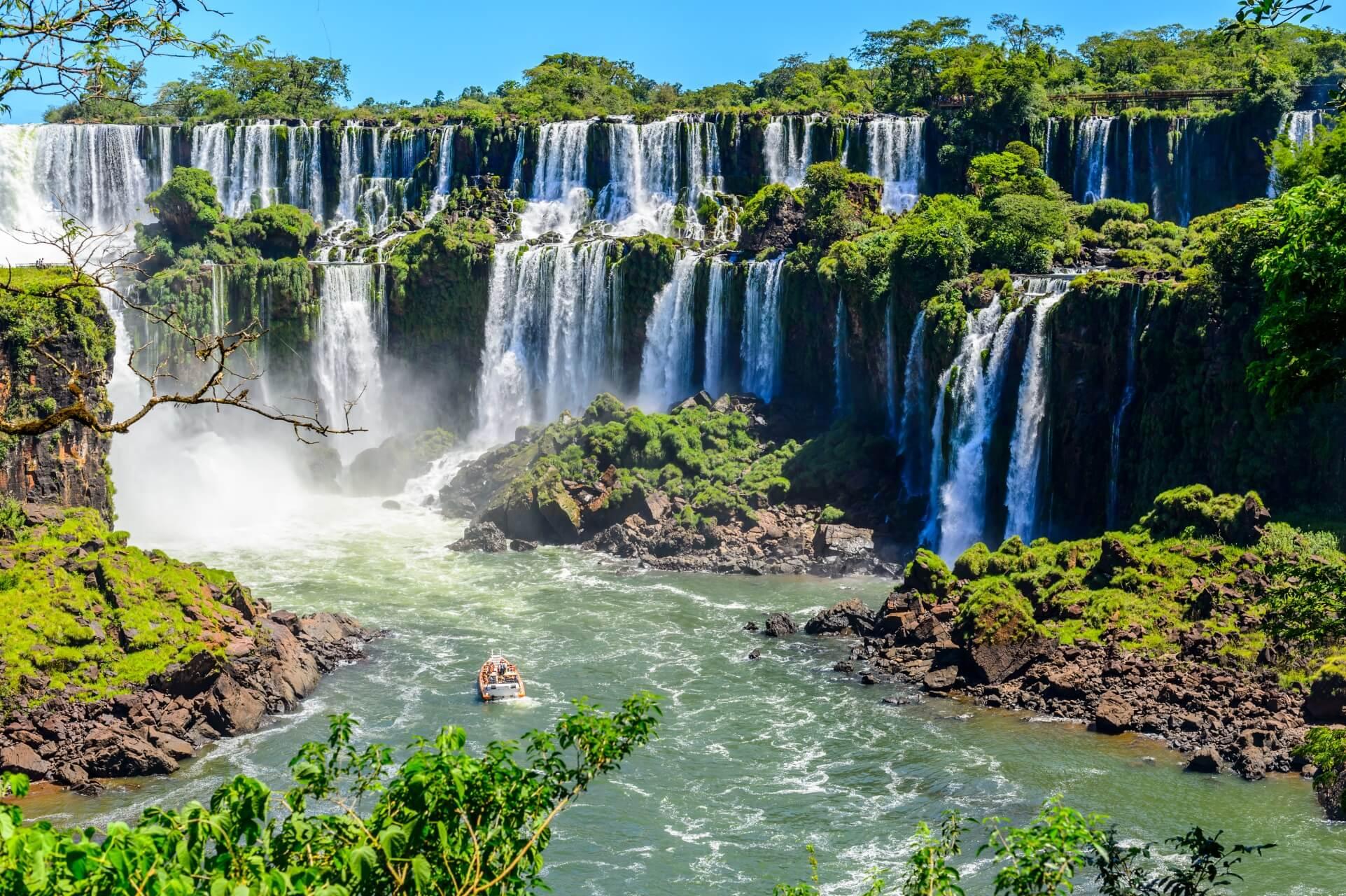 Cascatas majestosas de Iguazú caem de penhascos cobertos de vegetação. No primeiro plano, um barco navega em um amplo rio verde, cercado por vegetação exuberante e céu azul.