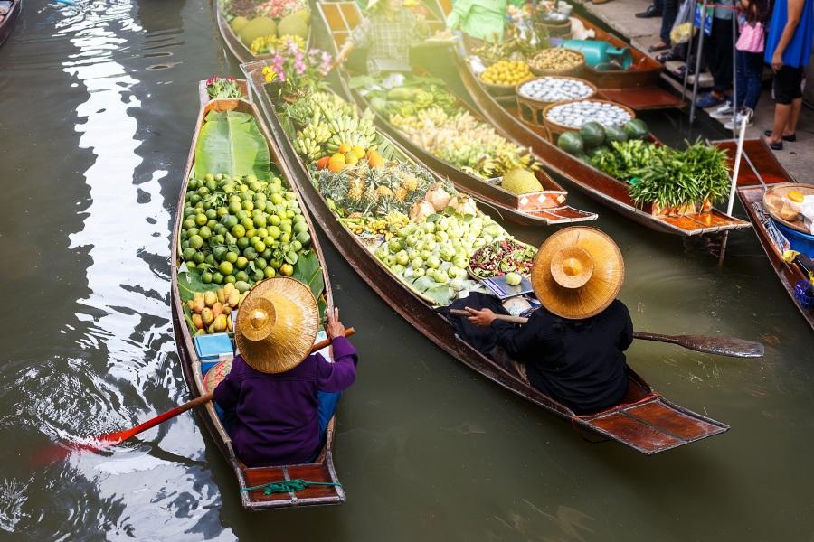 Boats at a floating market in Thailand are loaded with colorful fruits and vegetables. Two people in straw hats are rowing. One boat is on the left, two on the right, on green, calm water.
