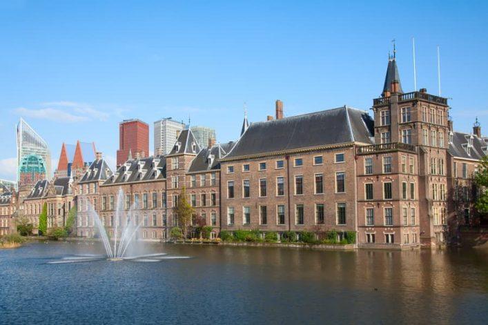Historic building with a brick facade and tower by the water, surrounded by modern architecture in the background. A fountain in the foreground adds movement to the water surface.