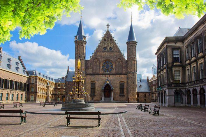 Historic building with distinctive towers in the background, a fountain with a golden statue in the foreground, surrounded by benches. Deciduous trees frame the top of the image, clear blue sky.