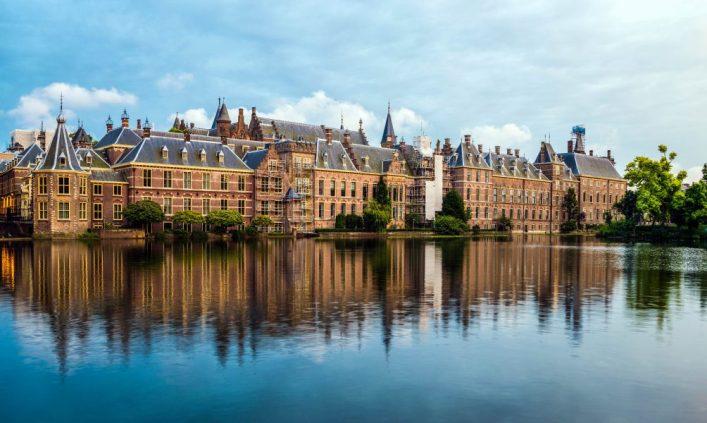 Historical buildings of the Binnenhof in The Hague, Netherlands, reflected in a calm pond. The sky is cloudy. Trees and bushes line the right edge.