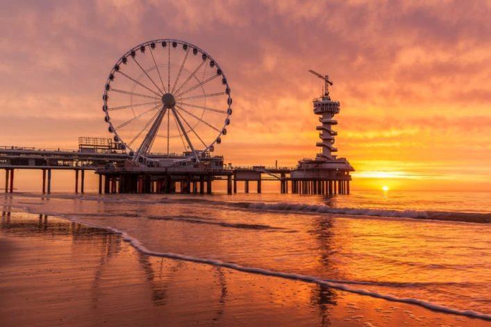 Giant Ferris wheel on a pier, flanked by a tower at sunset. The sky glows orange and reflects in the calm sea. Gentle waves on the sandy beach are in the foreground.