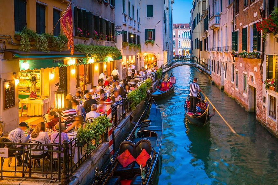 On the right side, a restaurant hugs the canal. Tables sit on a veranda above the water, it is already dusky, and the lights on the tables give the canal a romantic flair. A boat passes through the canal next to the restaurant.