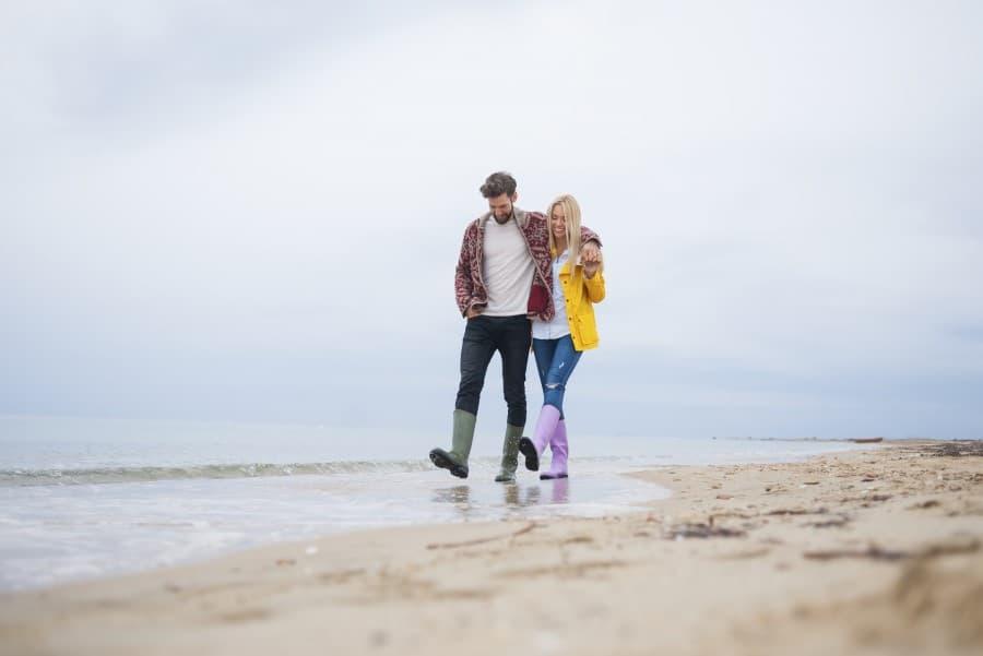 A young couple walks arm in arm along a beach. The weather is dreary and wet, and both individuals in the picture are wearing rubber boots; the woman is wearing a yellow rain jacket.