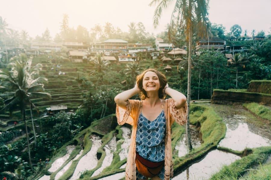 A young woman stands before the rice fields in Bali, holding her hair with both hands at the nape of her neck, tilting her head slightly back, and wearing a broad grin on her face.