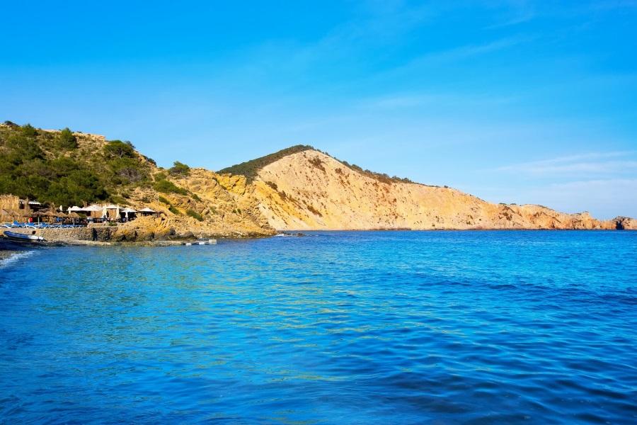 Clear blue sea with gentle waves in the foreground, rocky coast with green bushes on the left, golden cliffs in the background under a bright blue sky. Restaurant umbrellas on the left near the coast.