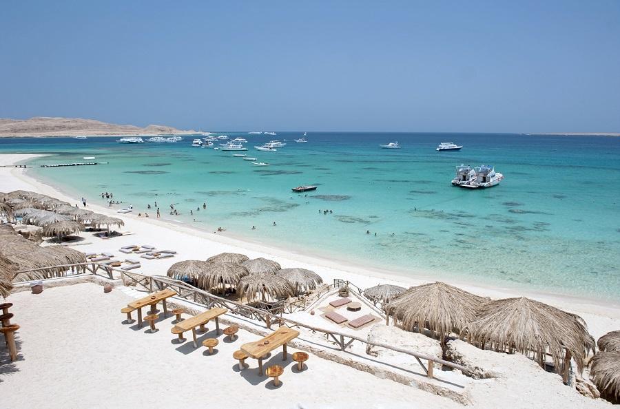 A bright sandy beach with straw umbrellas and wooden loungers stretches into the turquoise sea with several boats and snorkelers. In the background, rolling hills frame the wide bay, a perfect backdrop for popular cruise routes.