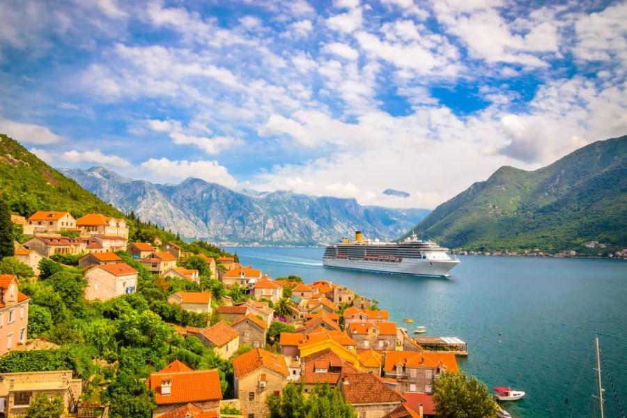 A cruise passes a picturesque coastal town with red roofs, green hills, and majestic mountains in the background. A cruise ship sails through the bay.