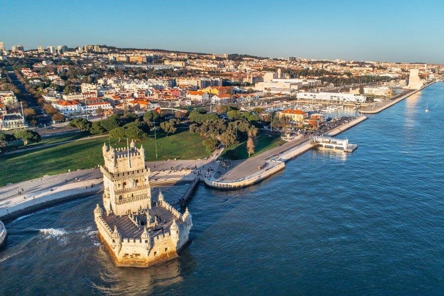Aerial view of the coastline of Lisbon with the famous Belém Tower in the foreground, surrounded by water. The well-kept waterfront stretches towards the city, which is known as a popular cruise destination.