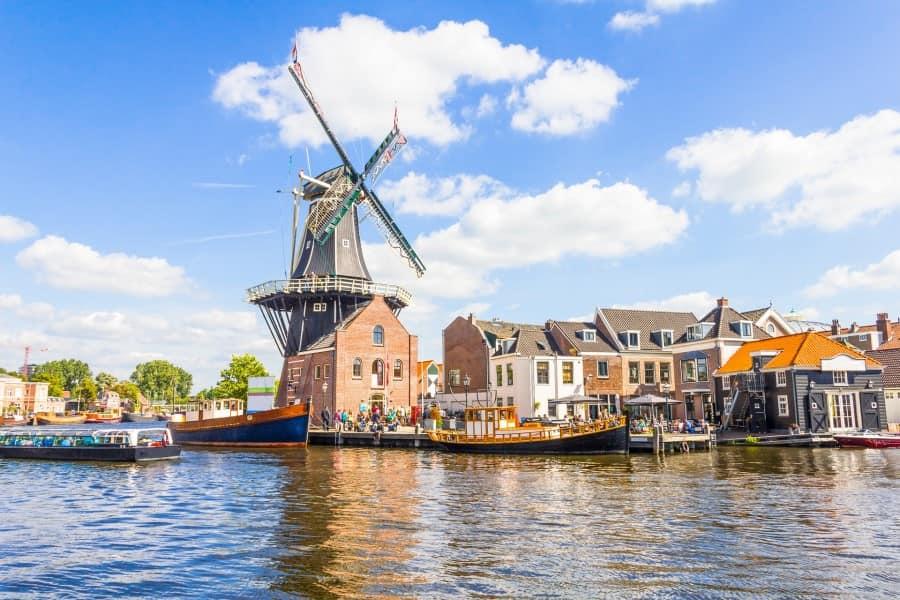 Traditional windmill by the riverbank in Haarlem, Netherlands, surrounded by historic houses in the sunshine. Blue water is traversed by boats that also navigate popular cruise routes.