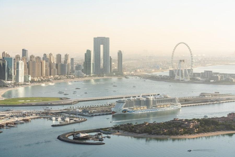 A modern cruise ship is docked in a major city's harbor with many skyscrapers on the left and the impressive Dubai Eye Ferris wheel on the right. Popular cruise routes pass through this region.