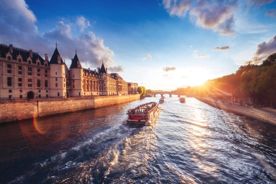 A cruise ship sails at sunset along the Seine, passing historic buildings on the left and green trees on the right. This image evokes memories of cruises through cities like Paris, Venice, or Amsterdam.