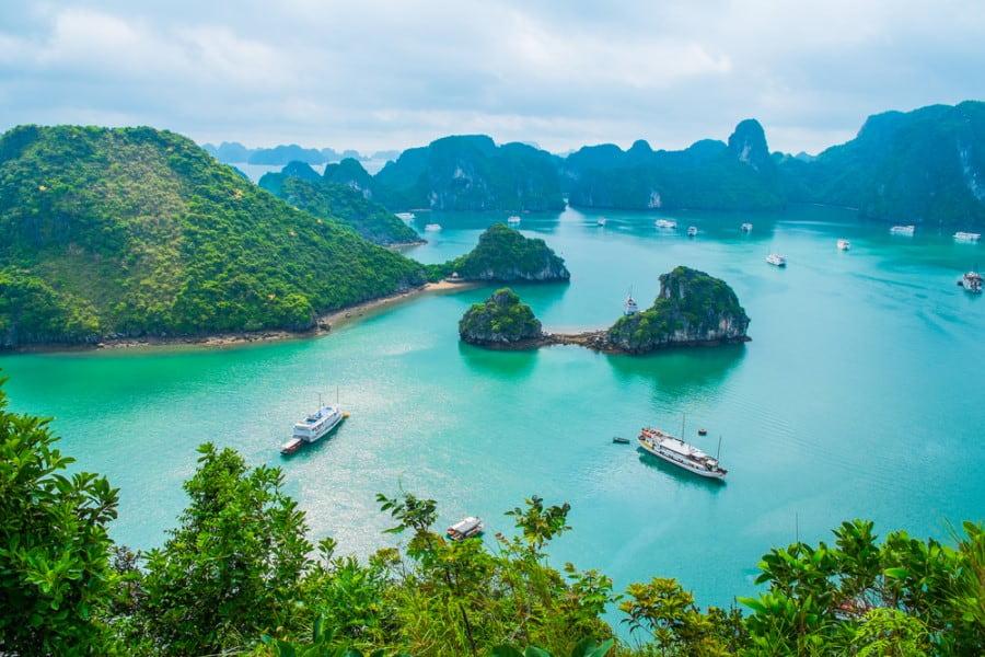 Turquoise waters in a bay with green, forested rock islands, typical of a popular cruise route. Several white boats and cruise ships sail between the islands.