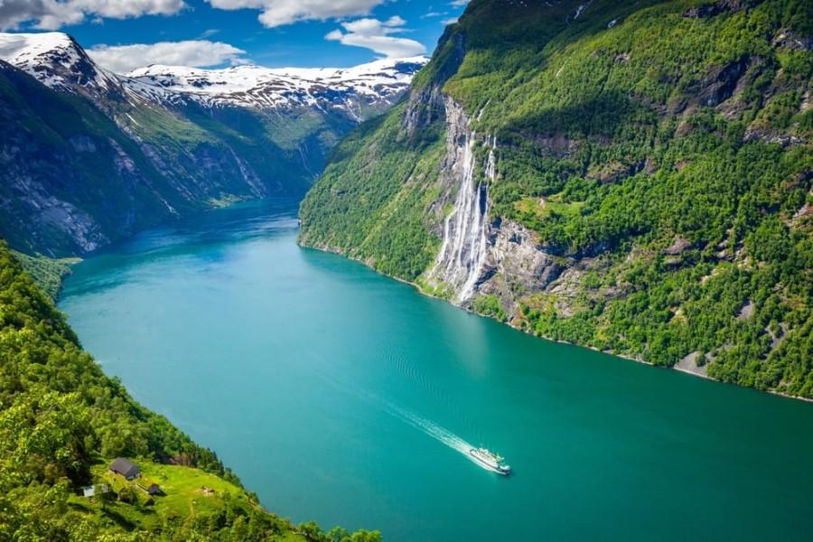 A cruise ship navigating through Geirangerfjord in Norway, a popular cruise route. To the right, an impressive waterfall cascades down the green, steep cliff face, with mountains with snow-covered peaks in the background.