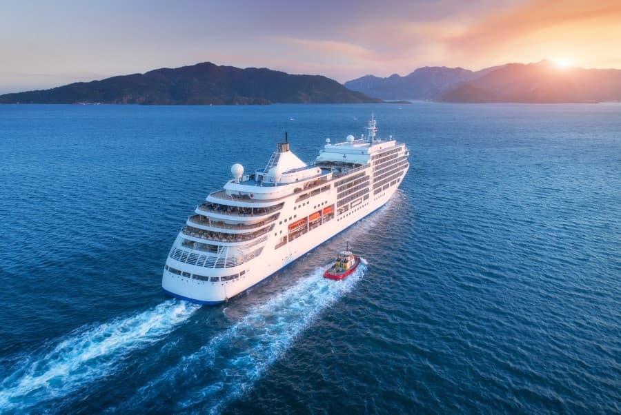 A cruise ship sailing in the Mediterranean at sunset, while an island chain is visible on the horizon. Popular cruise routes often travel along Italy, Greece, and Spain.
