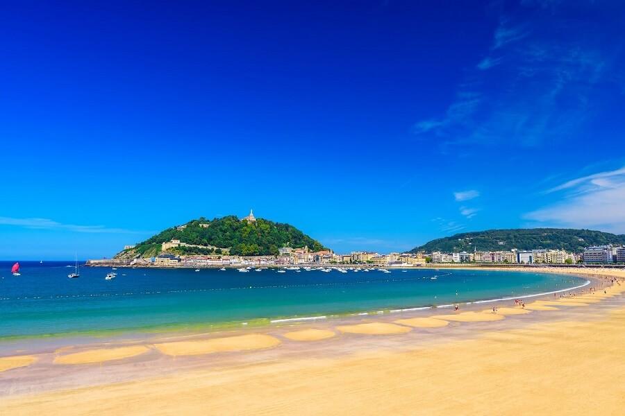 The white sand beach of La Concha in San Sebastián with turquoise water, behind it a hillside covered with green trees. Residential buildings to the left and right. Popular cruise routes pass here.