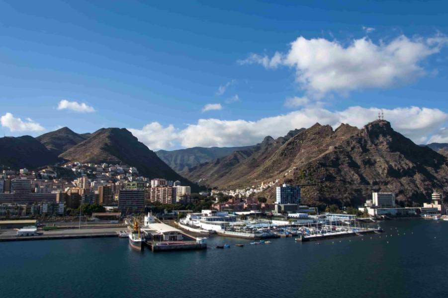 View of a busy harbor against green mountains, a blue sky, and scattered clouds. On the left are buildings of a city; on the right are docks and boats, popular cruise routes pass this breathtaking landscape.