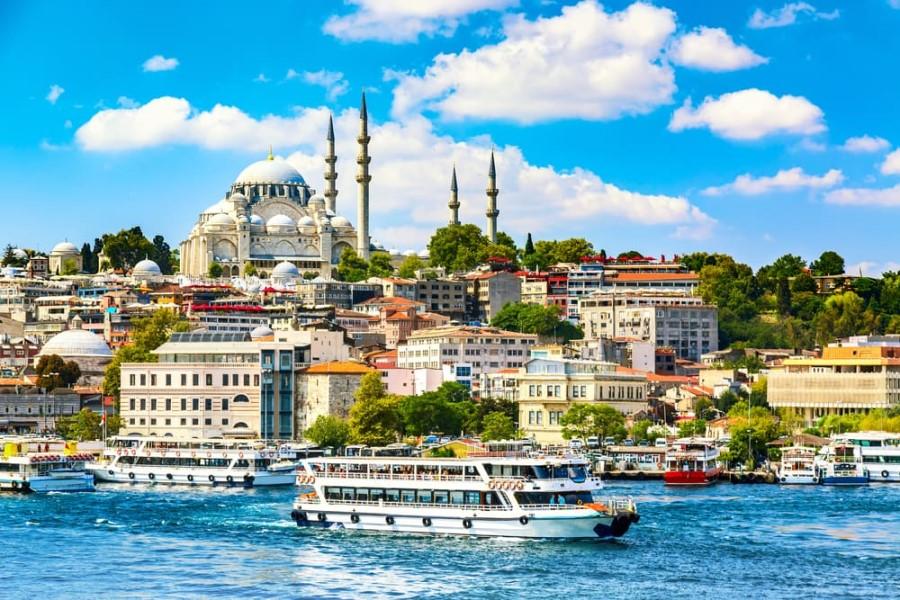 City skyline on the water in Istanbul with the magnificent Hagia Sophia in the background on a hill. In the harbor, several white boats are anchored and sailing, popular cruise routes. A bright blue sky with scattered clouds rounds off the scenery.