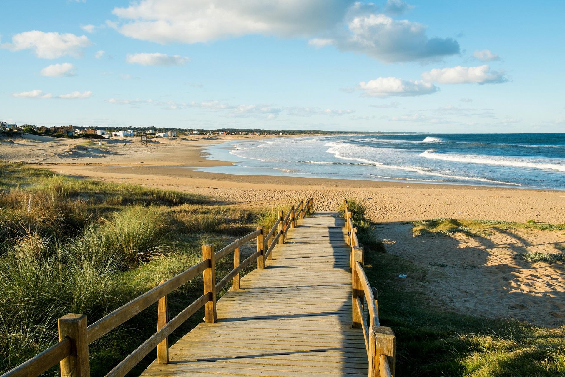 A wooden walkway leads over a sand dune landscape down to a wide sandy beach with gentle waves. In the background, the sea stretches under a blue sky.