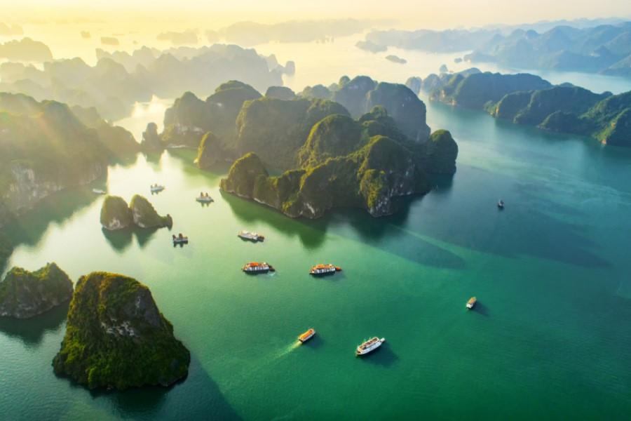 Several cruise ships sail through the turquoise Bay of Halong in Vietnam, passing forested limestone islands under a hazy sky. A popular destination on cruise routes.