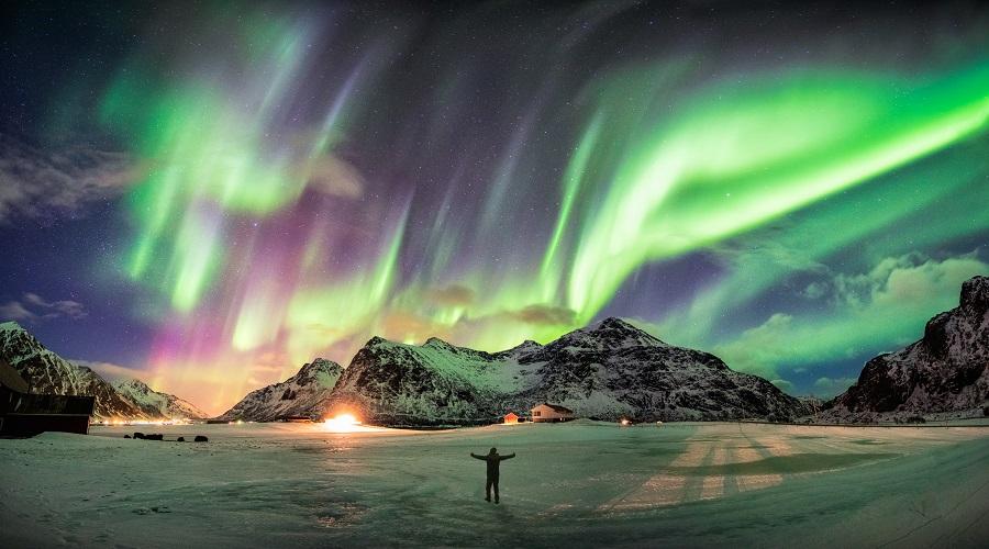 Colorful Northern Lights illuminate the night sky above snow-covered mountains and houses. A person stands with arms outstretched in a white snowy landscape, popular on cruise routes along the Norwegian coast.