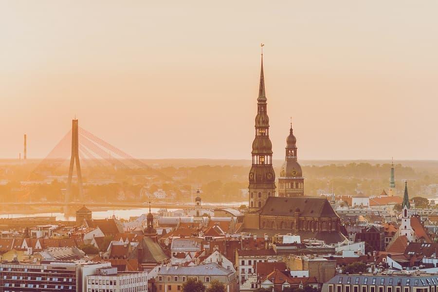 St. Peter's Church in Riga with its 132-meter-high tower overshadows the old town. In the background, the Vanšu Bridge and a gently glowing sunset can be seen. Popular cruise routes often lead here.