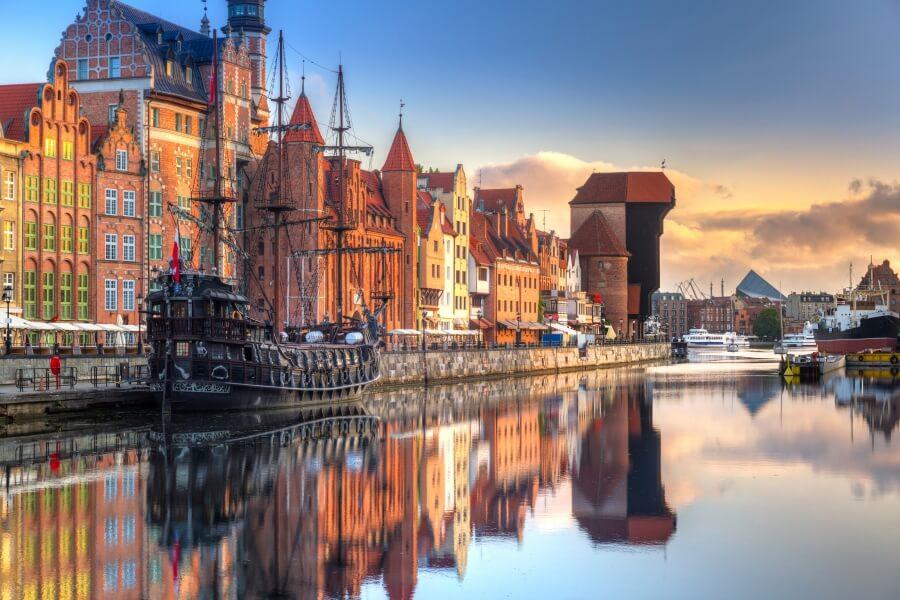 Colorful old town houses and a historic sailing ship reflect in the calm waters of the Motława in Gdansk, a popular cruise destination. In the background, the Crane and modern buildings rise.
