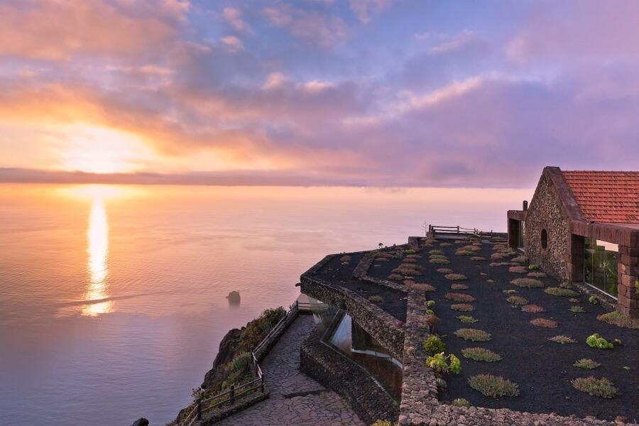 A sunset over the sea at a plant-covered terrace cliff on El Hierro, a popular destination on the cruise route of the Canary Islands. On the left is the sea, and on the right, a stone house with red tiles.