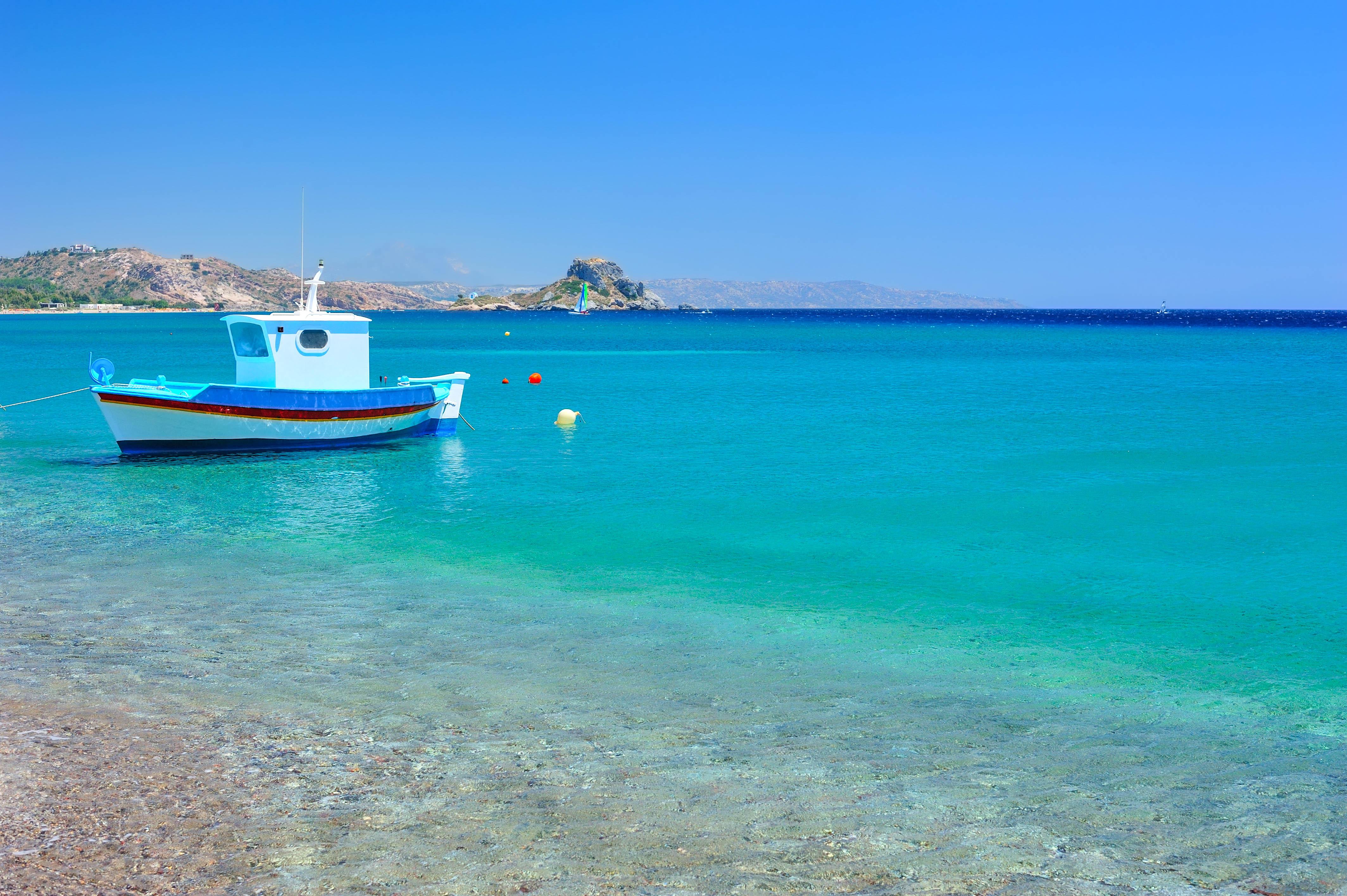 A turquoise Mediterranean sea with a small blue boat in the foreground, lying along popular cruise routes. In the background, a rocky island can be seen, with a bright blue sky above.