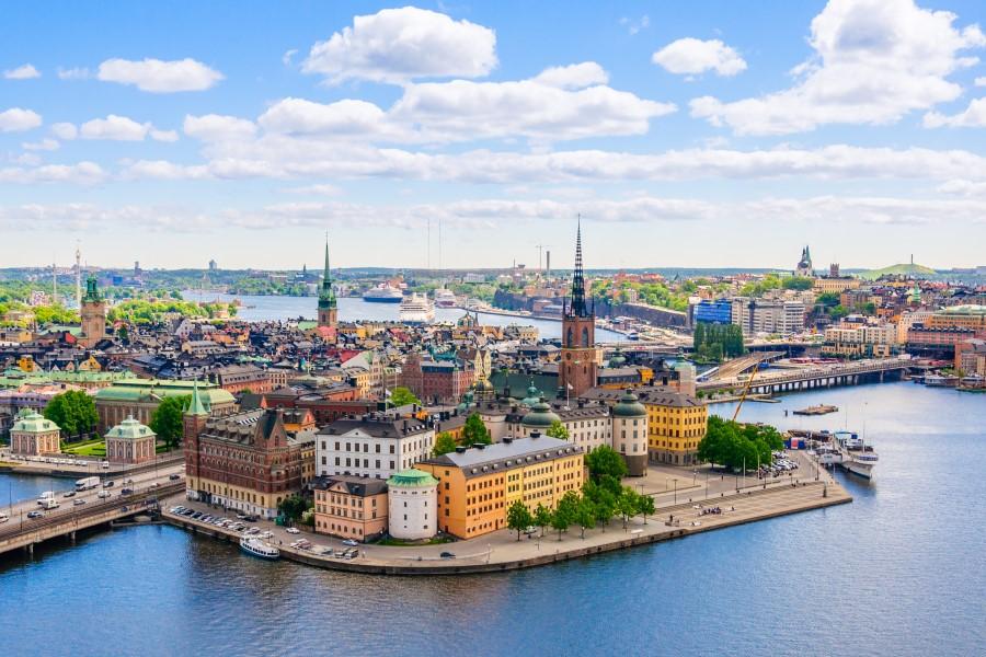 View of Stockholm with colorful historic houses and soaring church towers under the open sky. In the background, boats are seen. Riverbanks are visible on the right and left.