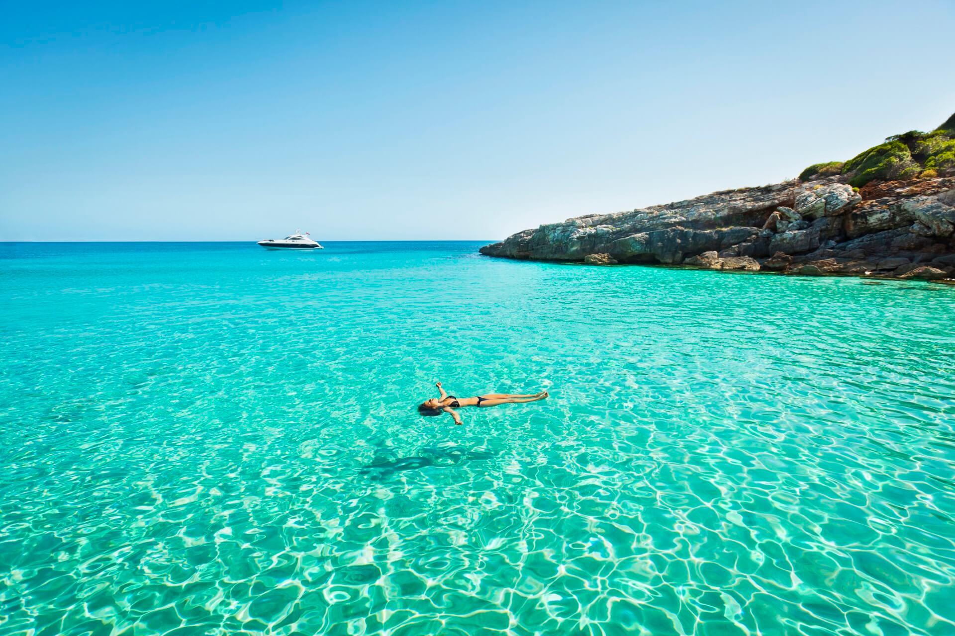 A woman swims leisurely in the clear turquoise Mediterranean near a rocky coast. In the background, a ship navigates a popular cruise route. The sun shines under a blue sky.