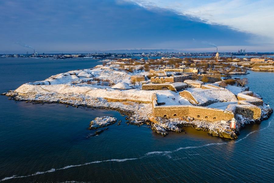 Aerial view of Suomenlinna in winter. The snow-covered fortress lies on an island, surrounded by blue sea, and in the background, the city of Helsinki with buildings is visible.