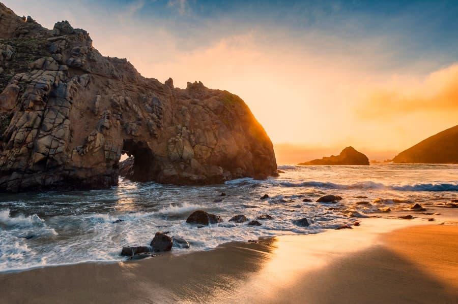 Sunset at Pfeiffer Beach. From the left edge of the image, a large rock juts into the frame, standing in the water. Behind it, the sun is setting, thus the image is predominantly yellow.