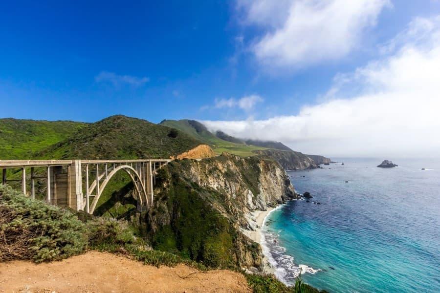 The view of the Bixby Bridge from a slight distance shows that the bridge is built in an arch shape. It stretches over the Pacific Ocean.