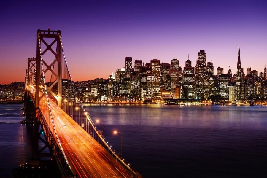 On the left side of the image, the Golden Gate Bridge spans over the water, glowing due to the cars driving on it, in a bright yellow-orange. In the background, the skyline of San Francisco can be seen, with tall buildings competing for height. The picture was taken at dusk.
