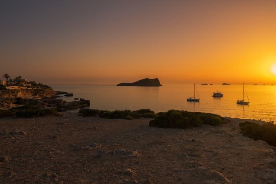 Sunset at Cala Comte in Ibiza. The sun sets at the right edge of the image, bathing the entire scene in a warm orange hue. Several boats are on the water, with houses and palm trees visible in the distance on the left side.