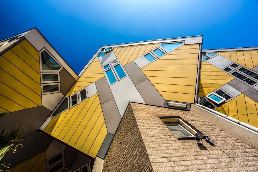 Yellow and white cube houses in Rotterdam, awkwardly standing on pillars. In the foreground is a brown wall, and the sky is bright blue.