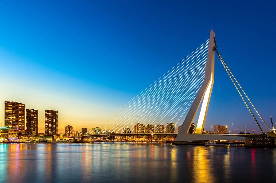 Erasmus Bridge in Rotterdam at dusk, featuring a distinctive slanted pylon and taut cables, with illuminated skyscrapers in the background and a clear, reflective water surface in the foreground.