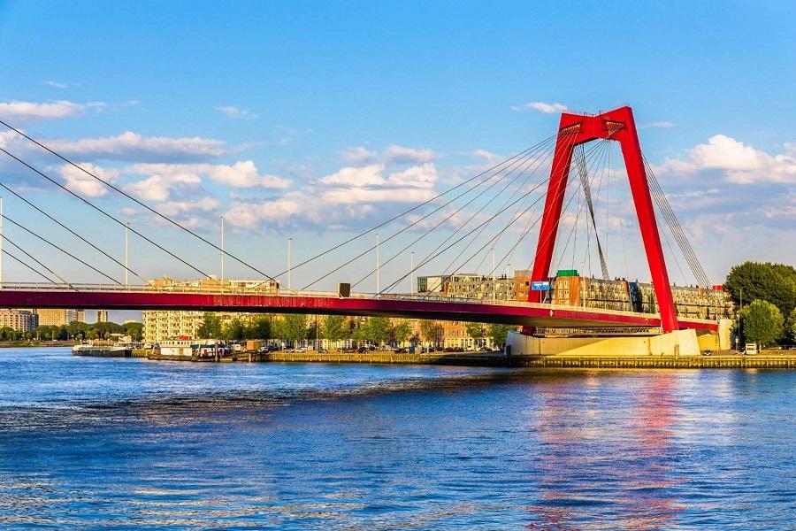 Red suspension bridge with distinctive beams and struts over a river; in the background, modern buildings and a blue sky with white clouds; the water reflects the sky.