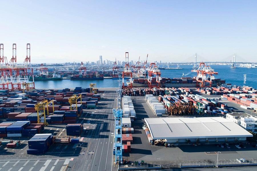 A sprawling port with numerous containers and cranes, with a skyline of tall buildings in the background. A large bridge spans the water on the right.