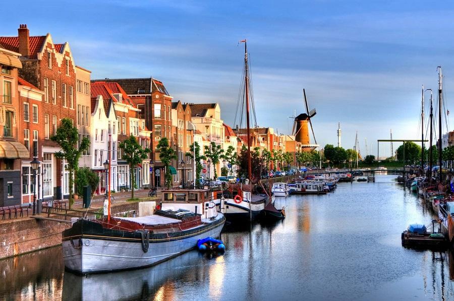 Sailboats are moored at the calm canal in Rotterdam. To the left are colorful old town houses, and to the right rises a black windmill against the blue sky. Trees line the shore.