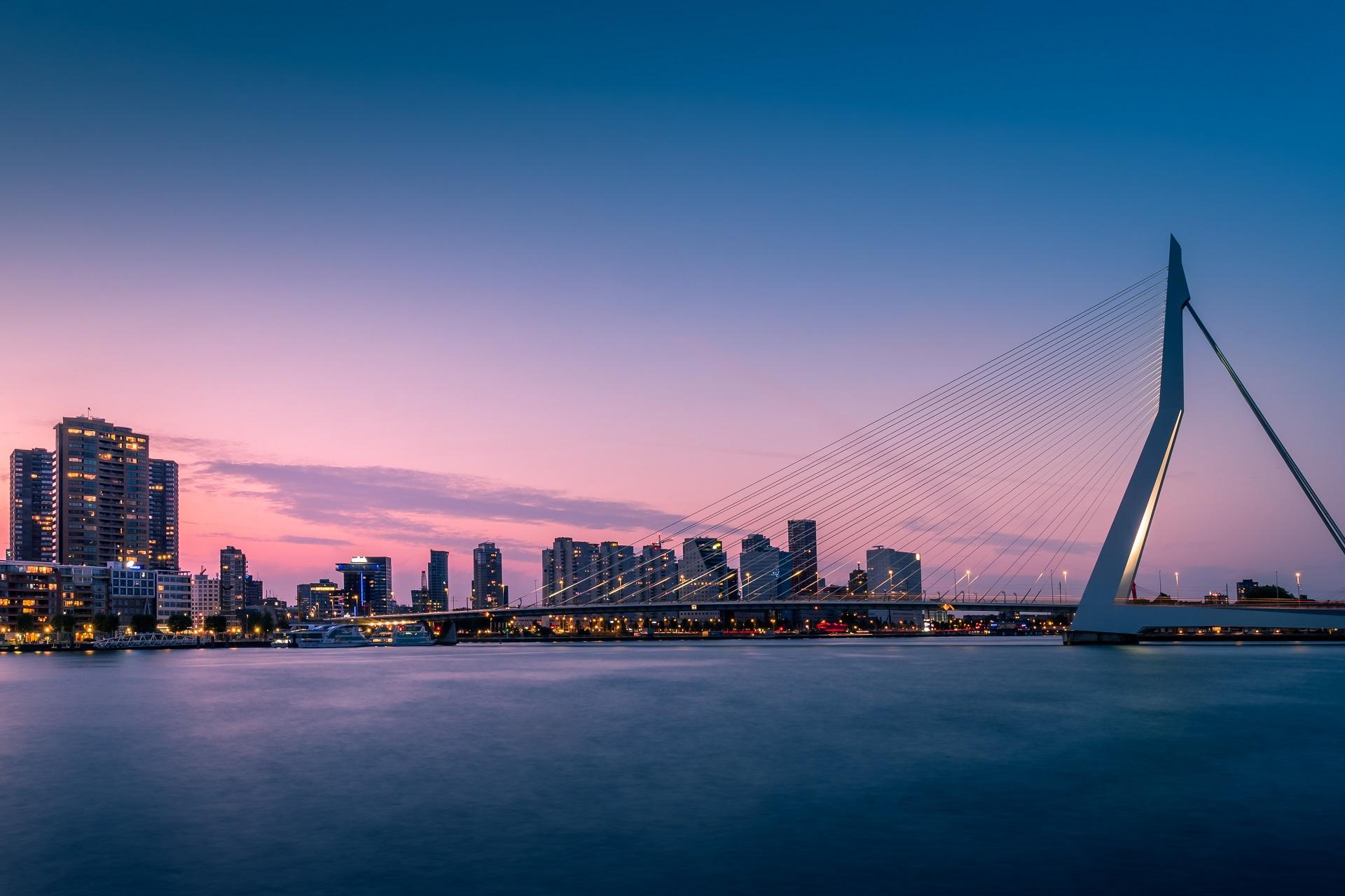 City view at sunset with modern skyscrapers on the horizon, a lit bridge on the right, and a calm river in the foreground reflecting the cityscape.