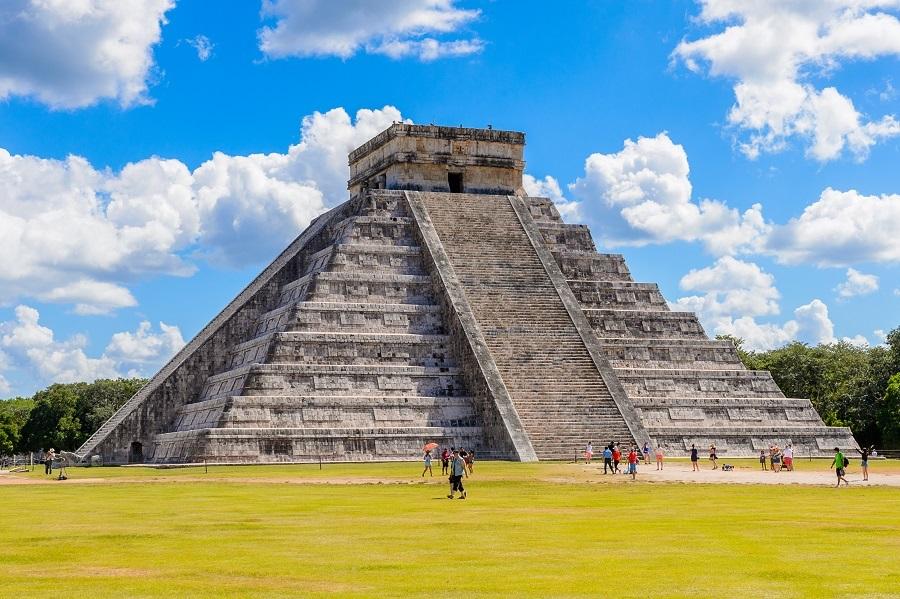 El Castillo Pyramid in Chichén Itzá against a blue sky with white clouds. Visitors are walking around the pyramid, which rises centrally on a green lawn.
