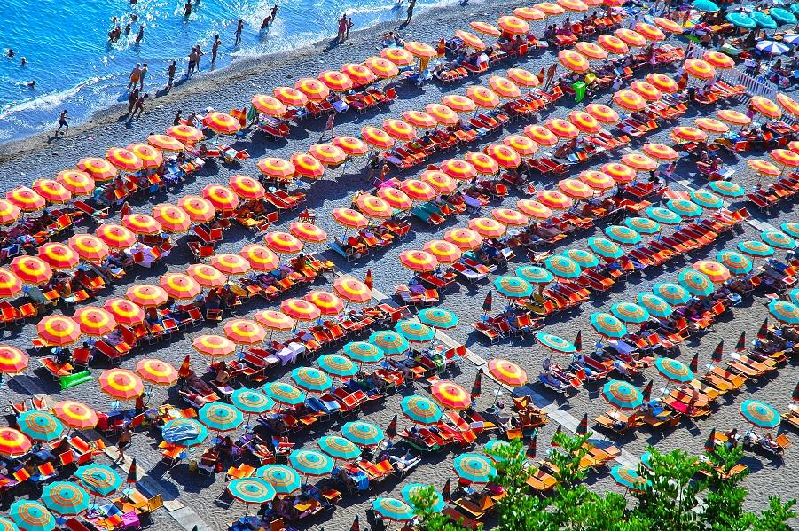 The beach of Positano offers numerous sun loungers. From the bird's-eye view, you can see green and orange red sun umbrellas lined up on the beach.