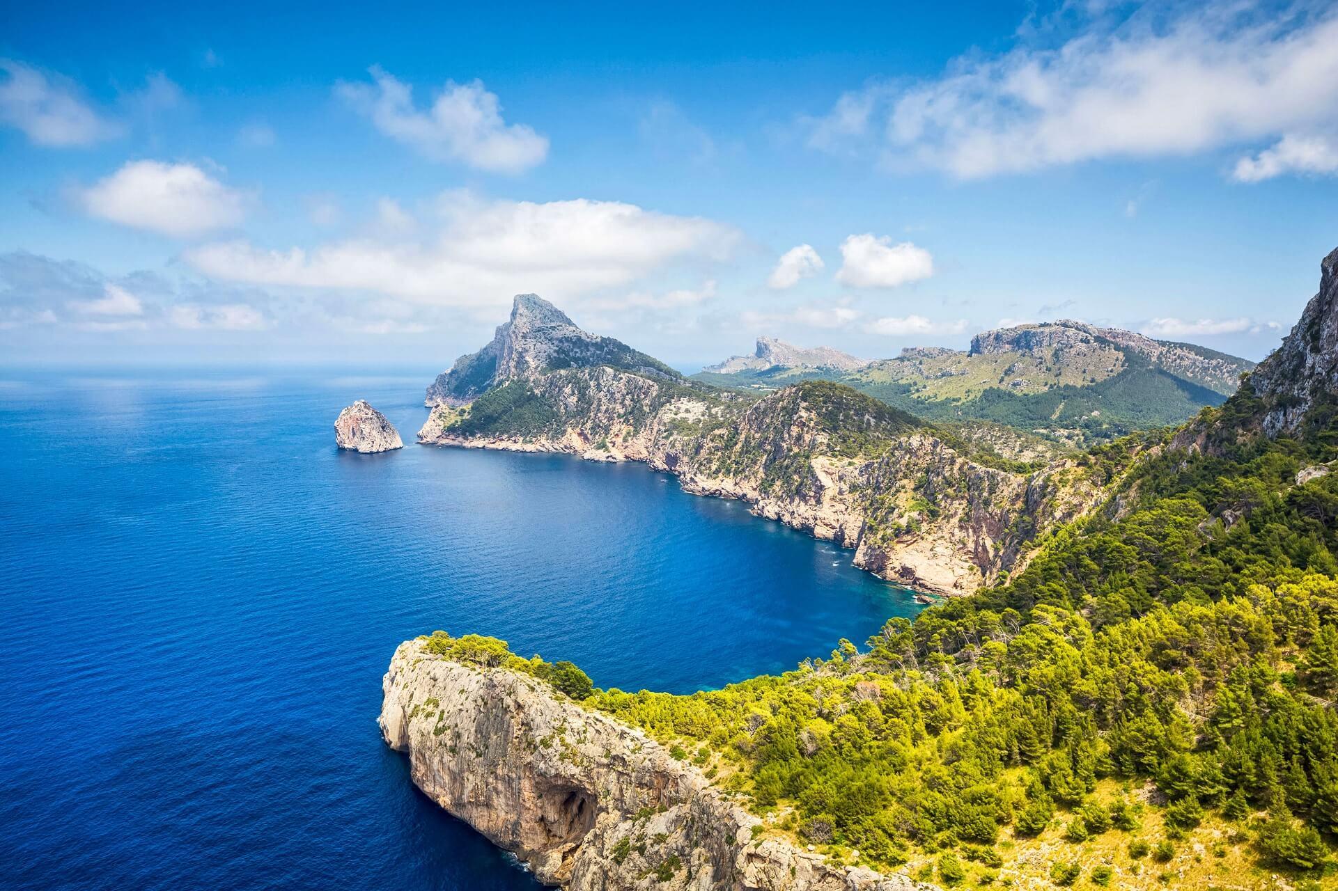 Steep, wooded cliffs rise into the deep blue sea, while the rugged coastline stretches into the distance. Above, a bright blue sky with white clouds.