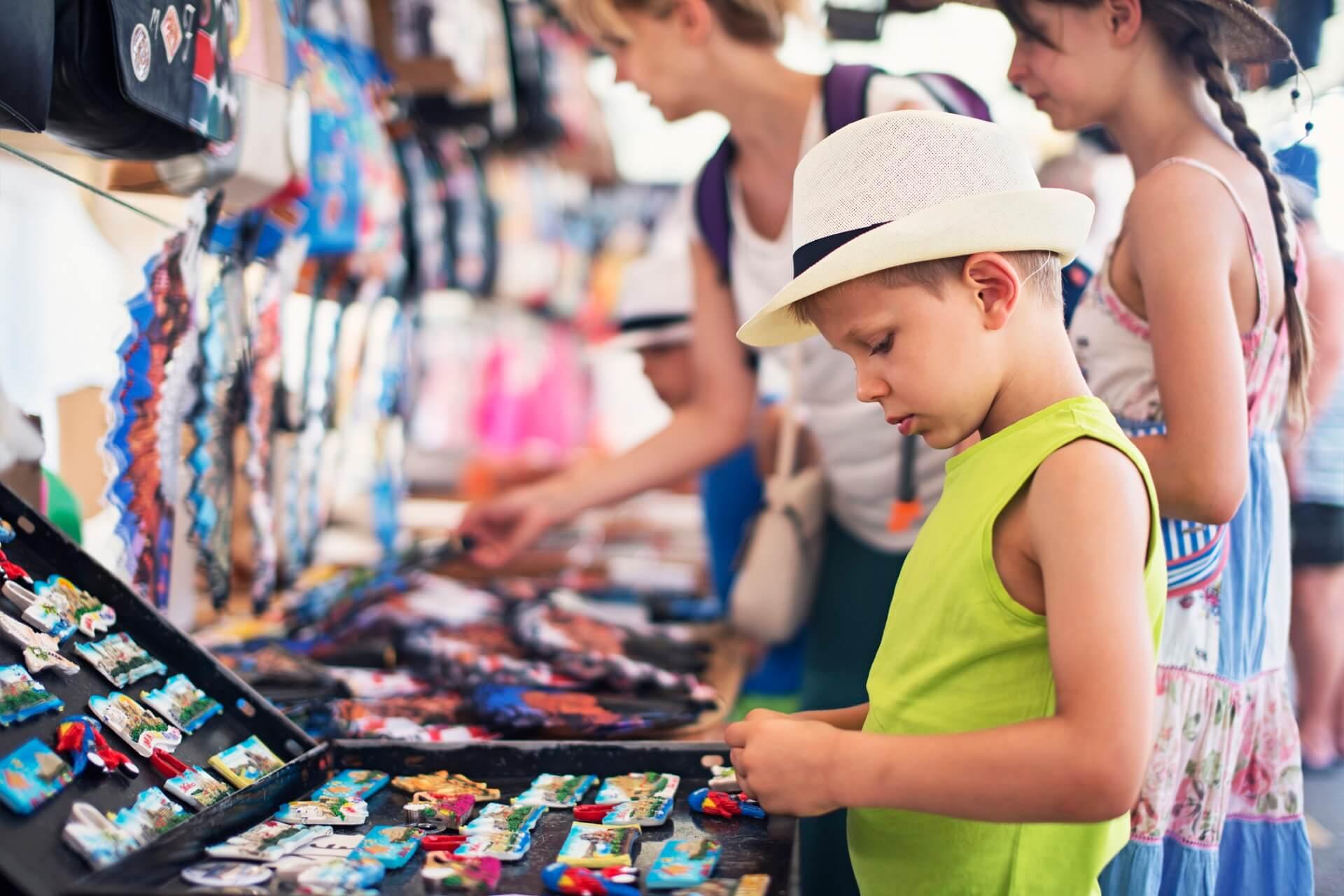 A young boy in the foreground with a white hat and green shirt contemplates colorful magnets at a market stand; in the background, other people are also examining the goods. On the left side, bags and souvenirs are displayed.