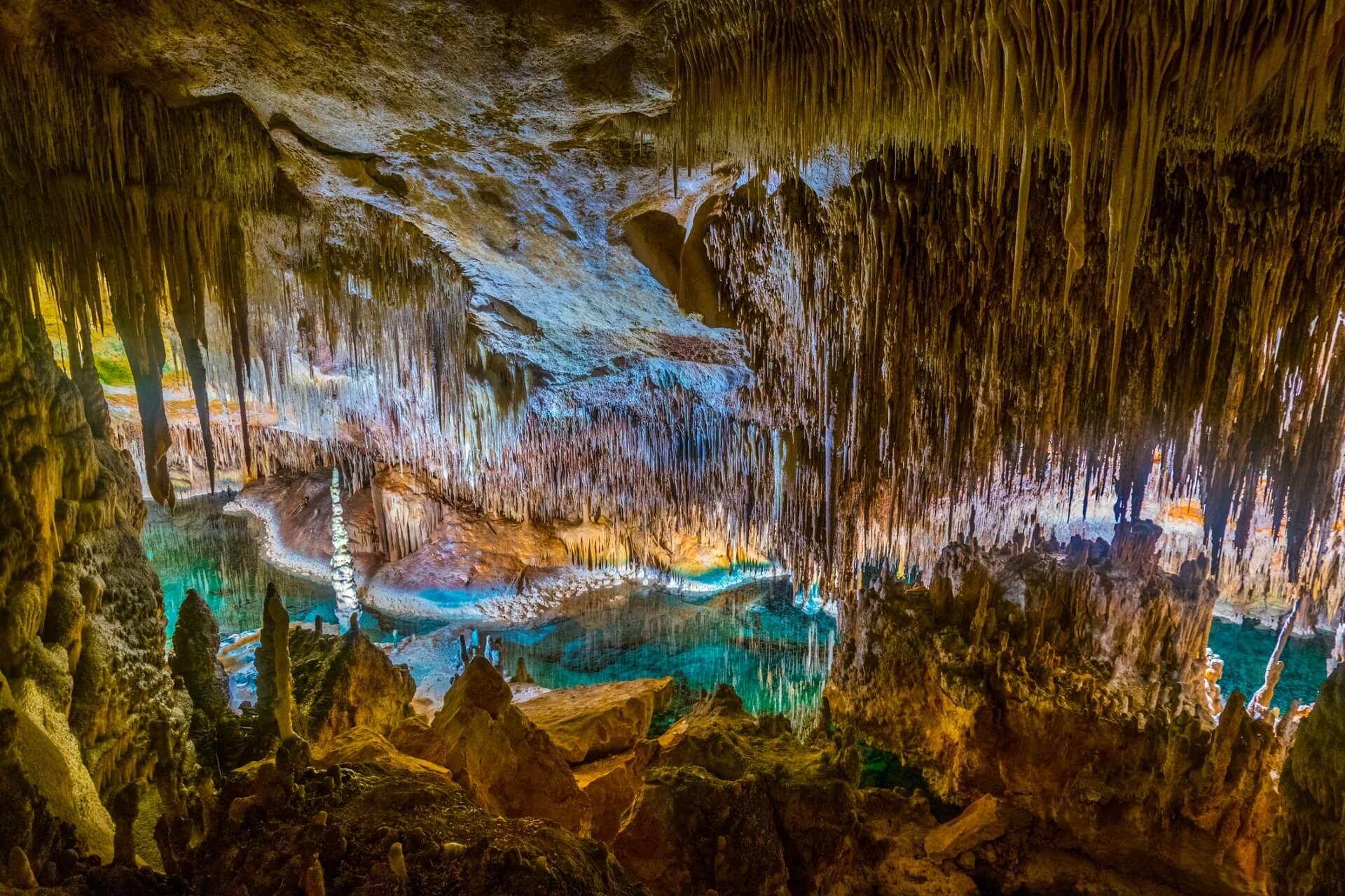 A dripstone cave with countless stalactites hanging from the ceiling, and stalagmites rising from the ground. In between, a clear, turquoise glowing underground lake reflects shimmering light.
