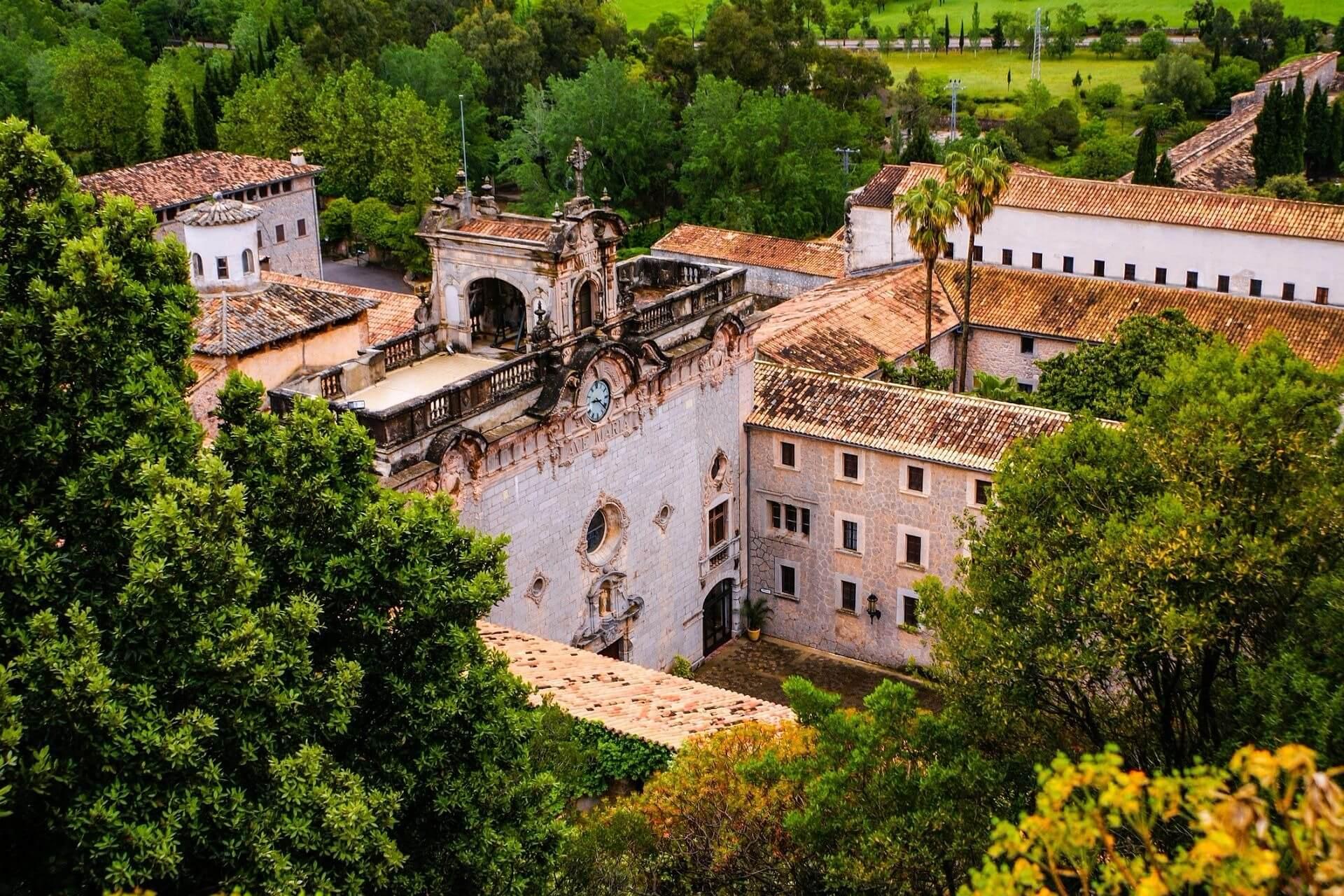 A historic building with stone facades and a red tile roof, nestled among lush green trees. In the foreground is a decorated entrance, with a wide green landscape in the background.