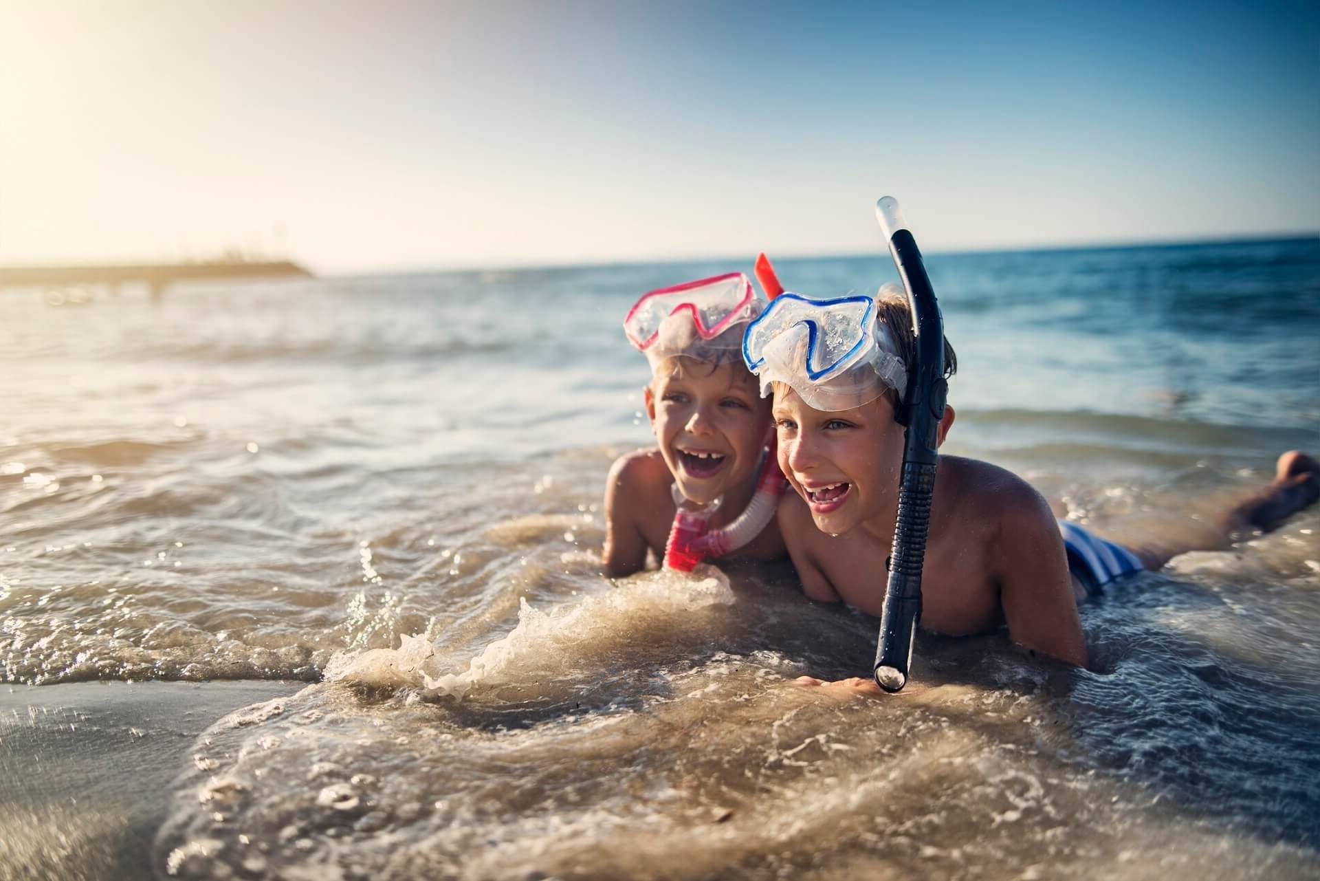 Two happy children with snorkeling gear play in the waves at the beach. The sun is shining, and the water splashes around them as they laugh in the shallow sea.