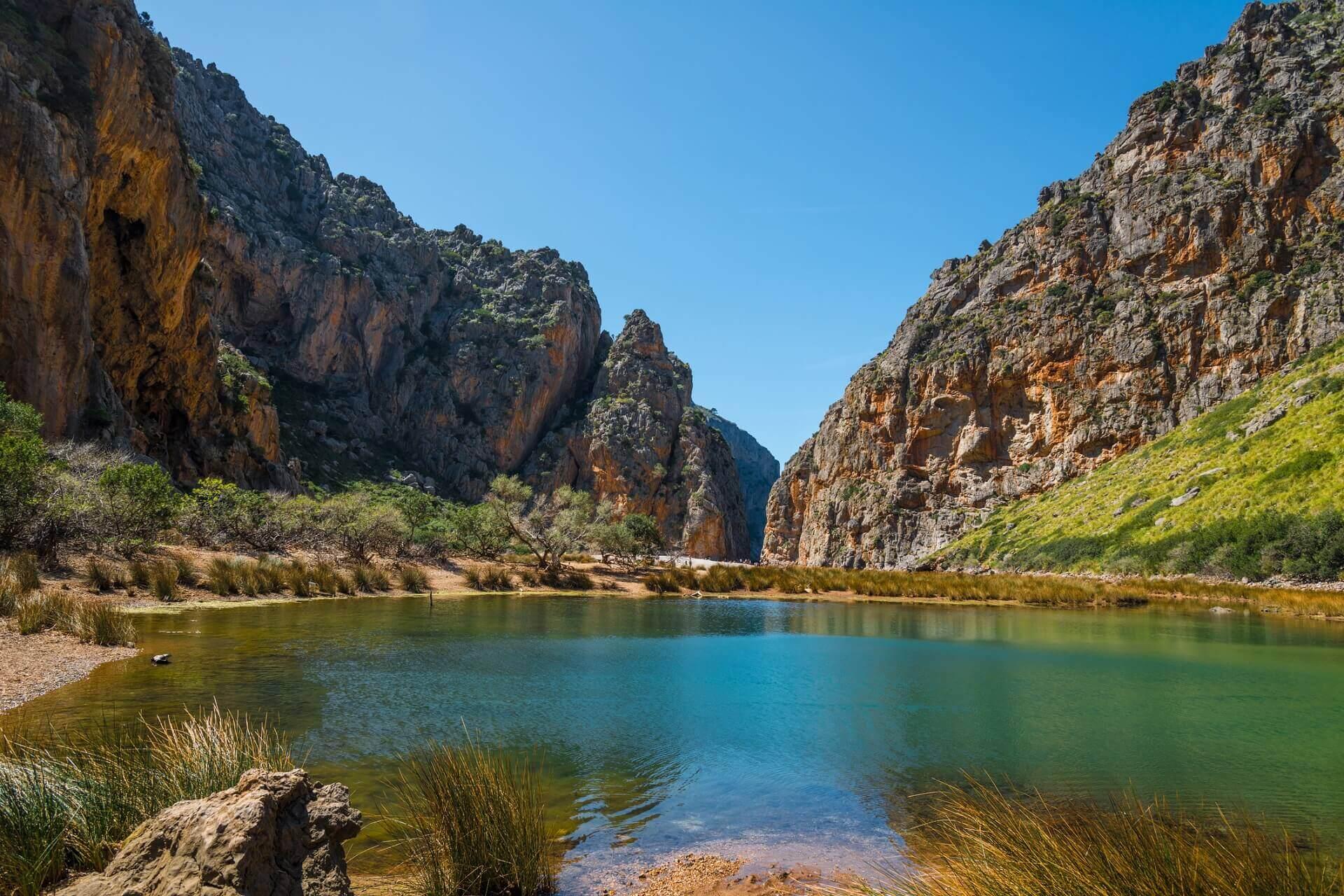 Clear, blue-green water of a lake in the foreground, surrounded by tall reeds. In the background, steep rocky cliffs rise under a blue sky and dense green bushes.